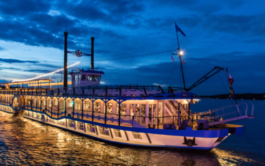 Ein Schiff fährt bei Anbruch der Dämmerung beleuchtet über einen Fluss.